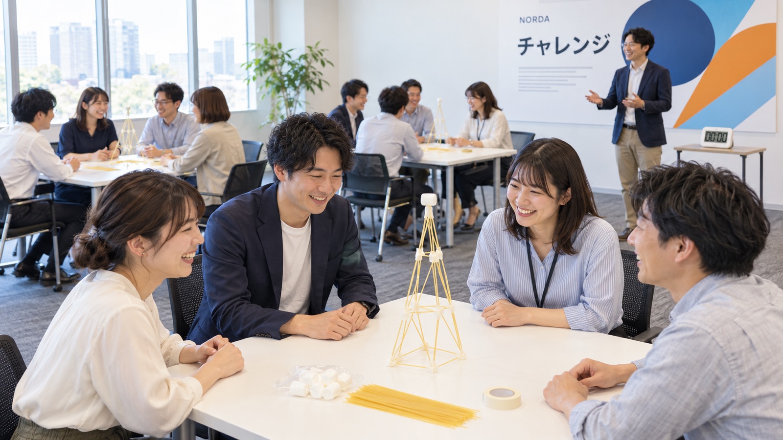 会議室で4つのチームが机を囲み、パスタとマシュマロでタワーを作る社内の懇親会の場面