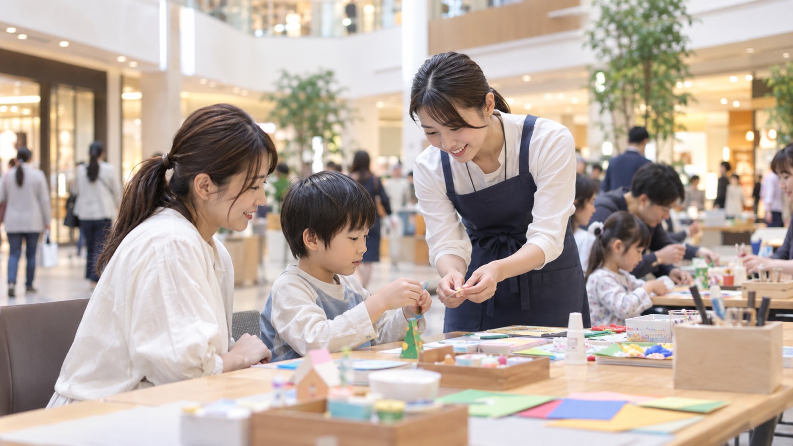 ショッピングモールの吹き抜け広場に並べた長机で、親子連れが座って工作のワークショップに参加している様子