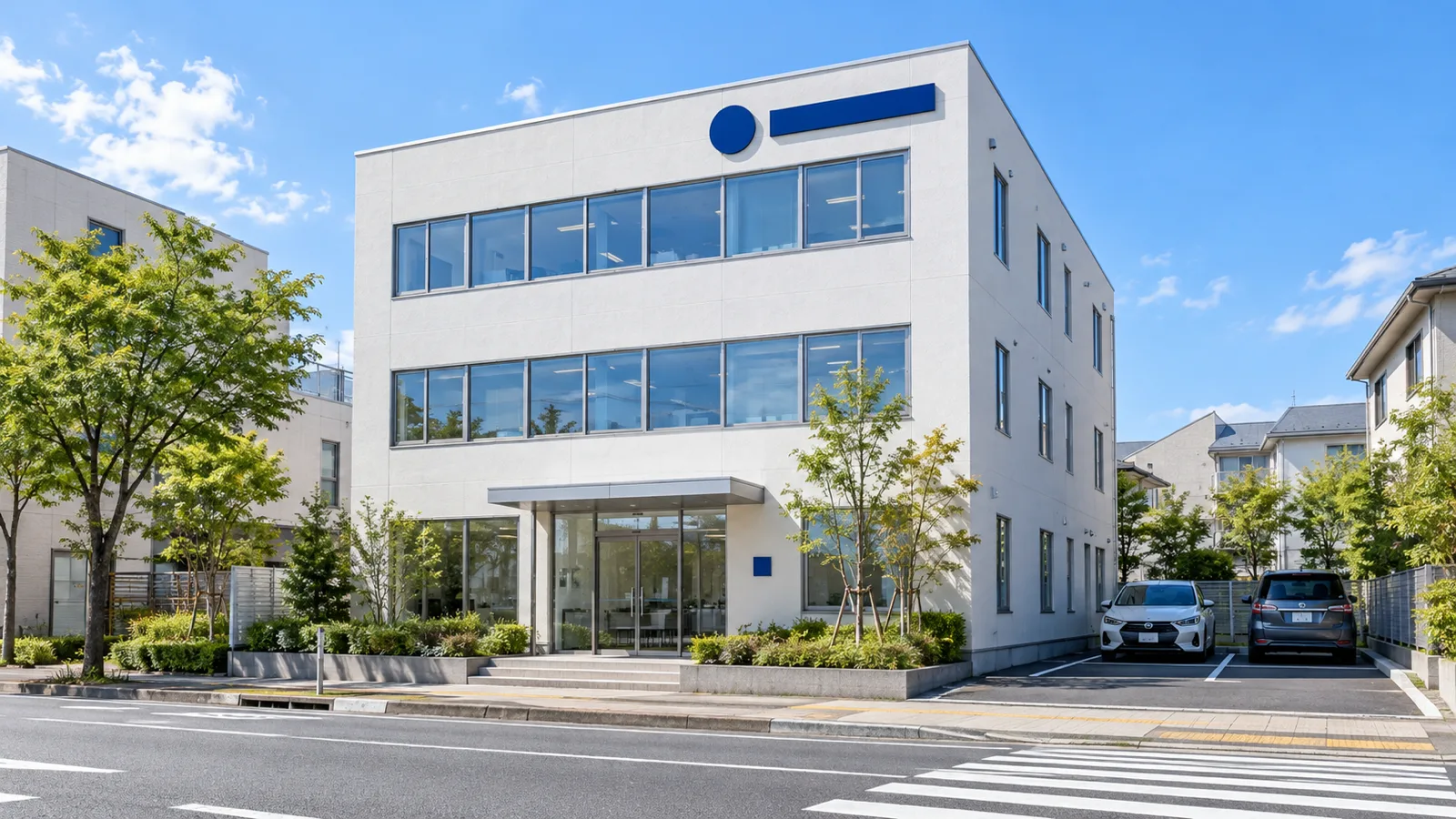 道路の向かい側の歩道から見た、白い外壁の3階建ての社屋の全景。3階の外壁の上部に紺色の無地の立体の帯と丸い形のマークが付き、1階の入口の横に小さな無地のプレートが見える様子