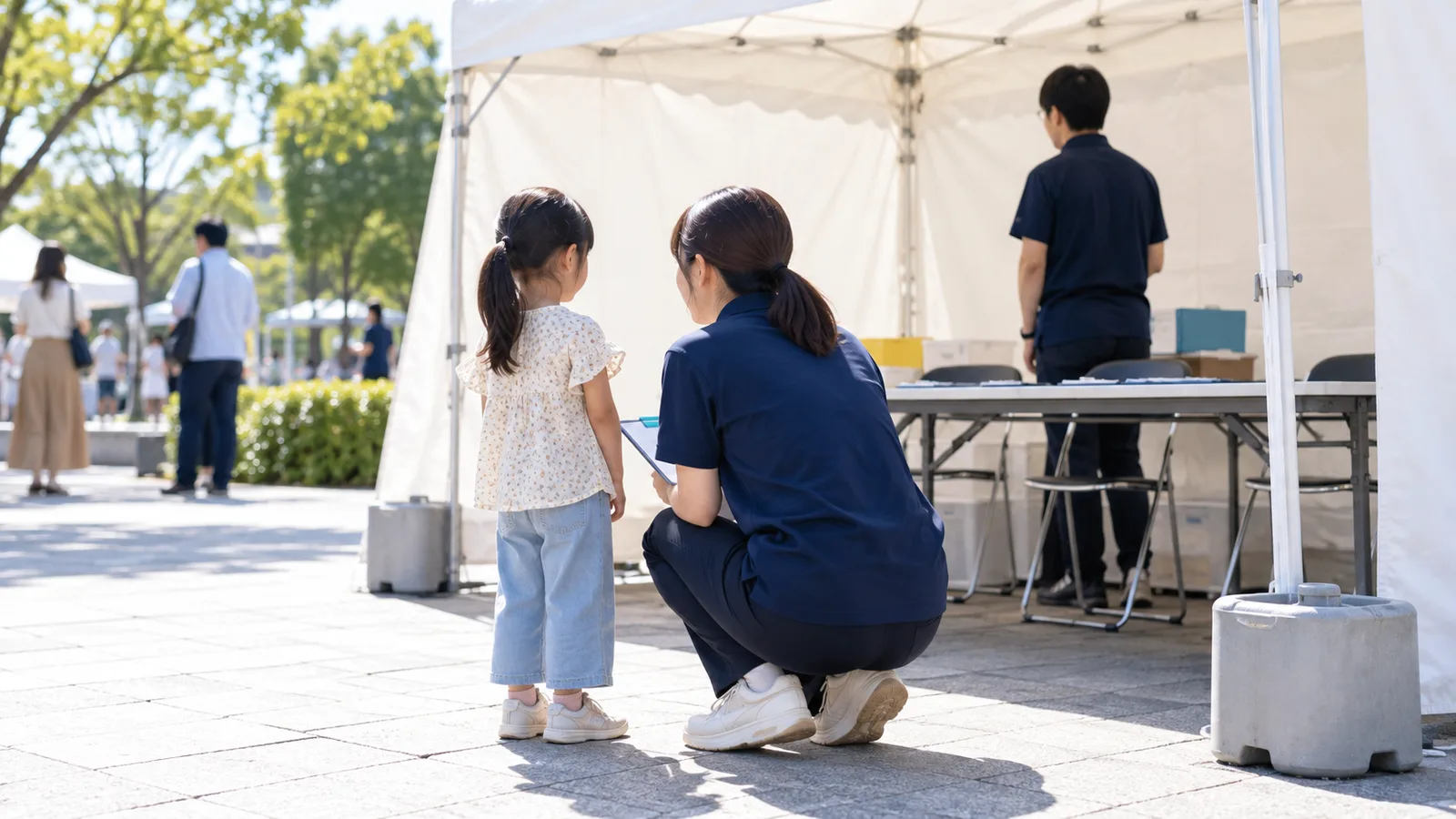 屋外イベントの本部のテントの前で、紺の無地のポロシャツのスタッフが、しゃがんで小さな子どもと目の高さを合わせて話を聞いている様子を、少し離れた斜め後ろから写した写真。子どもとスタッフの顔は写っていない