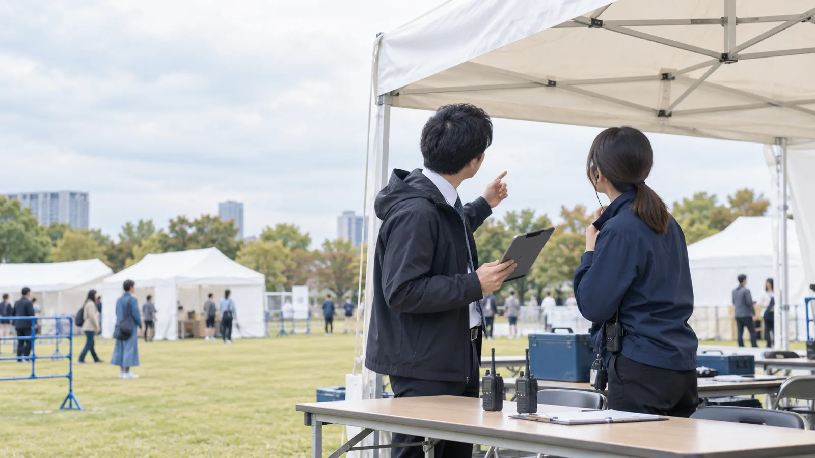屋外のイベント会場の白い本部テントの下で、2人の担当者がタブレットの画面を見ながら、曇り空のほうを見上げて話している様子を、テントの外から少し離れて写した写真。タブレットの画面は無地で、テントや机にも文字は無い