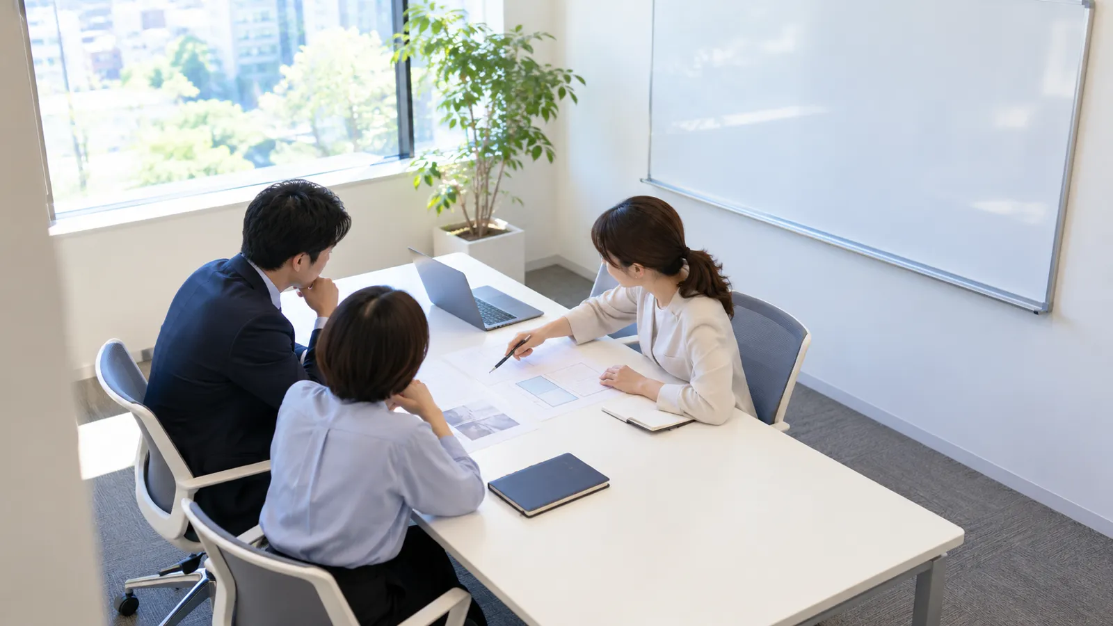小さな会議室で、社員3人が机を囲み、ノートパソコンと紙の資料を前に話し合っている様子を、部屋の隅から少し離れて写した写真。ホワイトボードは無地で、どこにも文字は無い。人物の顔は横顔か後ろ姿で、はっきりとは写っていない