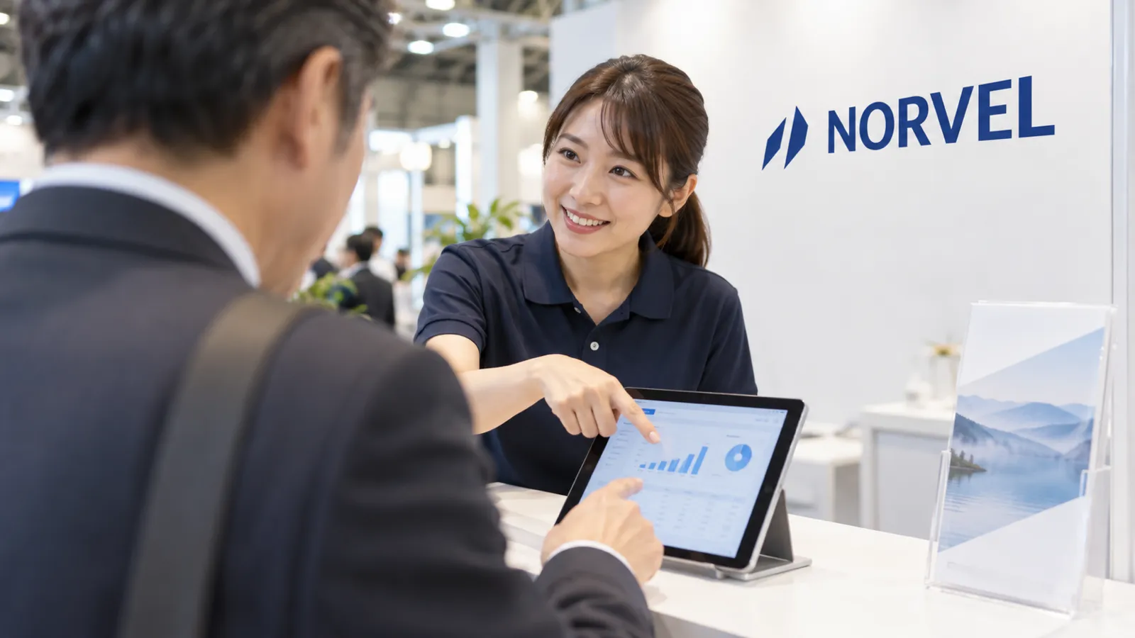 展示会ブースのカウンターで、来場者の男性がタブレットのデモ画面を自分の指で操作し、横に立つスタッフが画面を指しながら説明している様子を、来場者の肩越しに写した写真