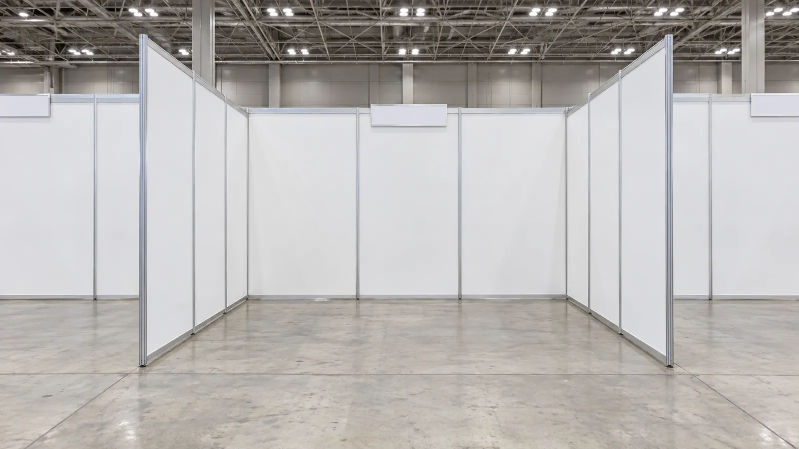 展示会場のホールで、白いシステムパネルの壁だけが立ち、床にはカーペットも什器もない3m×3mほどの空の小間を、通路の正面から大人の目の高さで写した写真。人物は写っていない