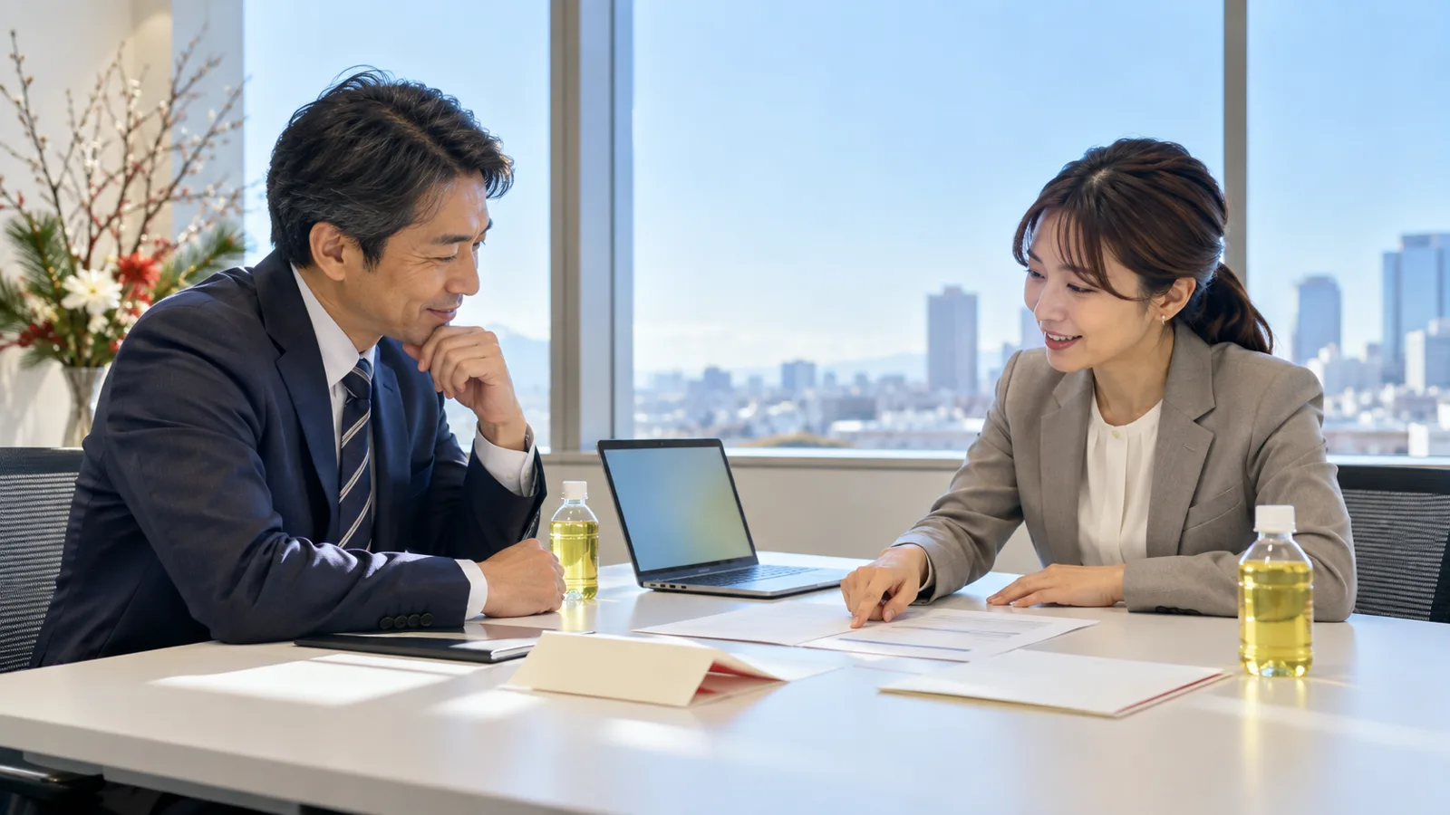 会社の会議室で、スーツ姿の男女2人が、テーブルに広げた前年の案内状と名簿の紙を指さしながら話し合っている様子を、横から写した写真