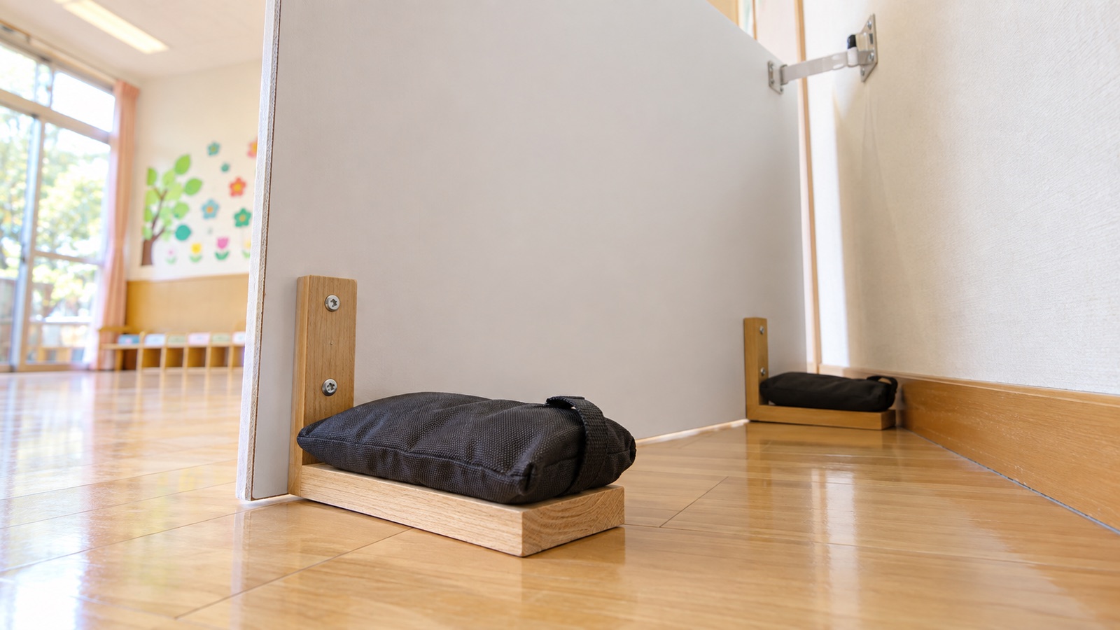 保育園の壁際に立てた顔はめパネルの裏側。脚の部分に重りを載せ、パネルの上部を壁の金具に留めている様子を、低い位置から写した接写