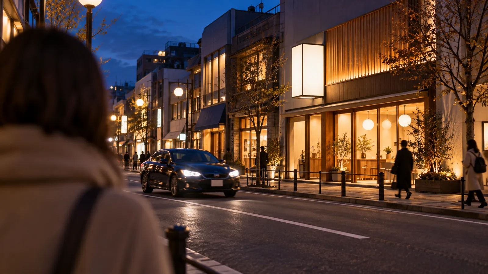 夕方の商店街で、道路の向かい側の歩道に立つコートの人の後ろ姿の肩越しに、向かいの店先の光る看板を見ている様子を写した写真。看板は無地で、通りには車が1台走っている。人物の顔は写っていない