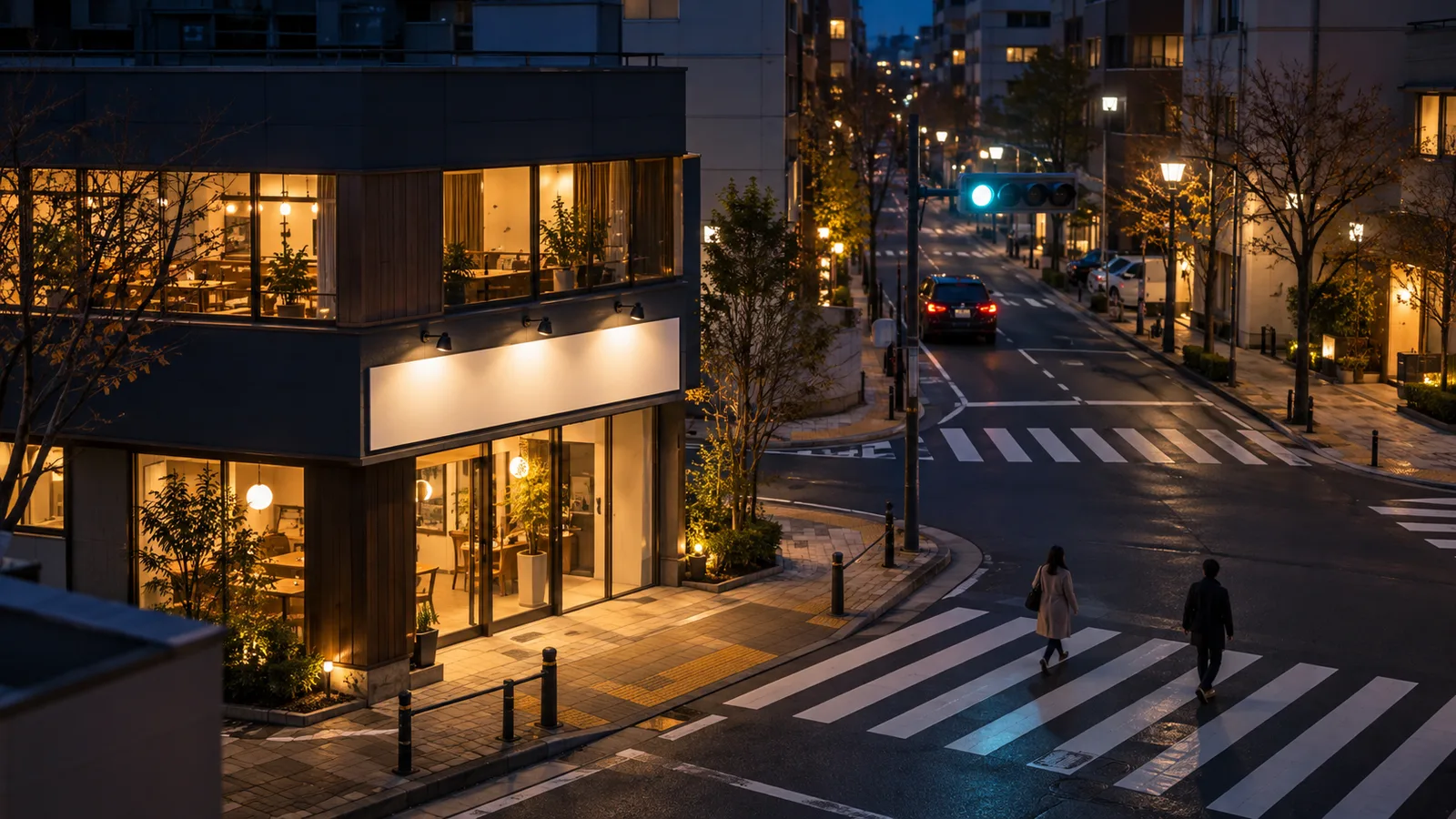 夜の交差点の角にある店舗の外壁の看板を、通りの向かいの建物の2階ほどの高さから見下ろして写した写真。看板は落ち着いた白い光で照らされ、交差点の奥に信号機が小さく見える。看板に文字は無く、人物は小さく写っている