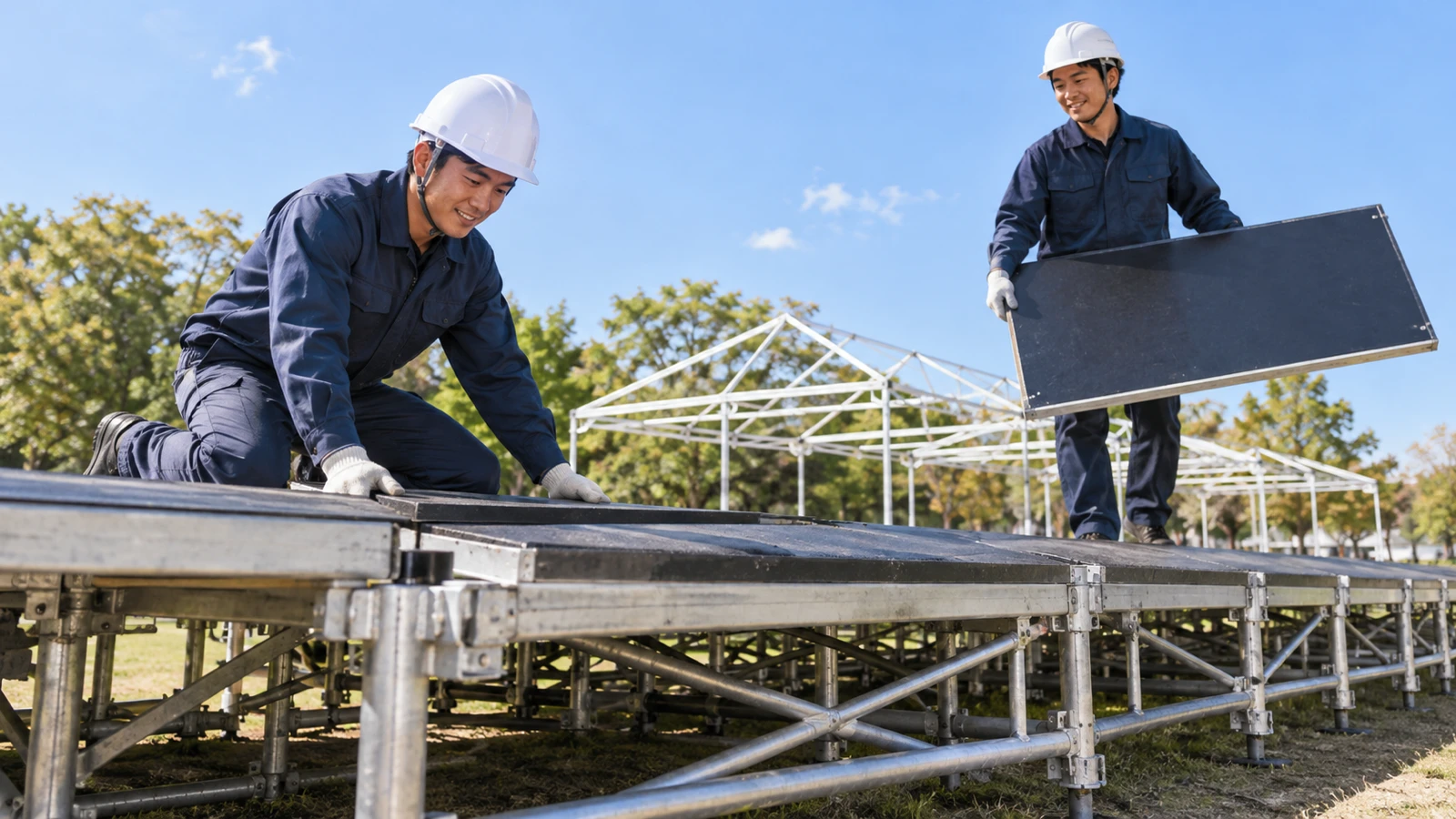 日中の公園の広場で、作業着の施工スタッフ2人が、組み上げ途中の仮設ステージの骨組みに床の板を据え付けている様子を、地面に近い低い位置から見上げるように写した写真。背後には設営前のテントの骨組みと青空が見える