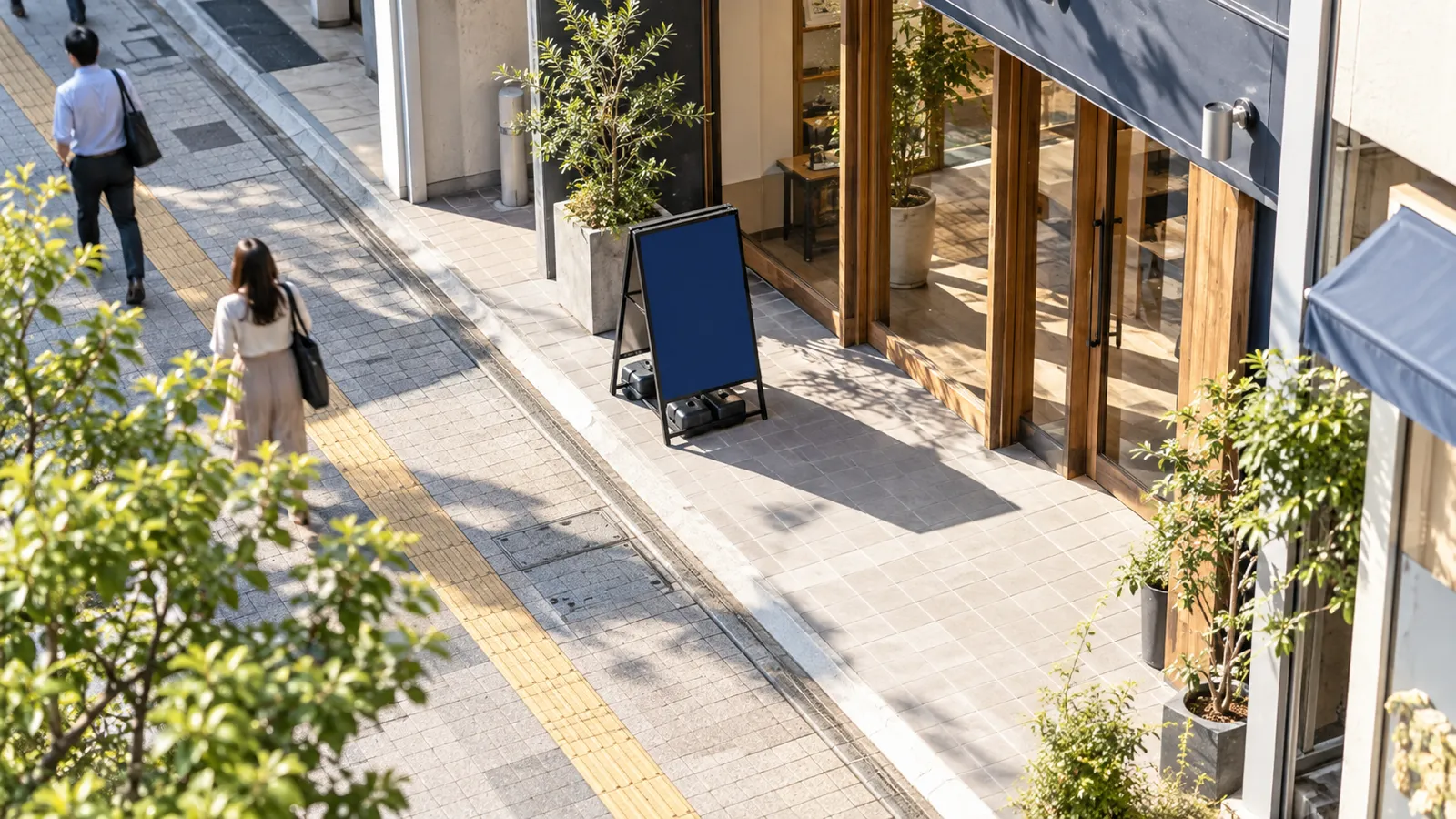 晴れた昼の店先で、店の敷地の中の入口の脇に無地のスタンド看板が置かれ、その先に歩道と点字ブロックが続いている様子を、2階ほどの高さから見下ろして写した写真。看板に文字は無く、歩道を歩く人が小さく写っている