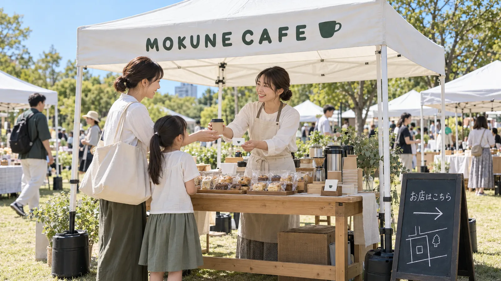 公園のマルシェで、カフェのテントのブースの店主が、母親と女の子に紙カップを渡しながらショップカードを差し出している様子。テントの横に「お店はこちら」と書いた黒板の看板が立っている