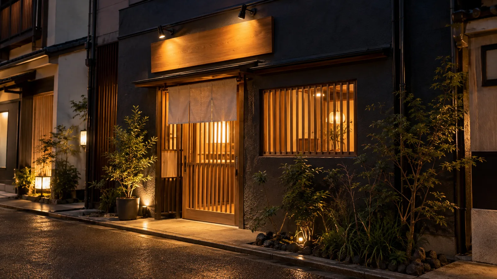 夜の路地に面した飲食店の店先を、少し離れた正面から写した写真。壁に付いた無地の木の看板が上から小さなスポットライトで照らされ、入口の格子戸から暖かい色の店内の明かりがこぼれている。周りは落ち着いた明るさで、人物は写っていない