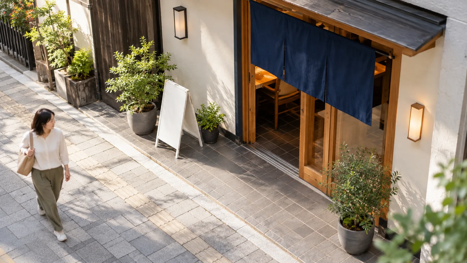 昼の路面店の入口を、少し高い位置から見下ろすように写した写真。A型の置き看板と鉢植えが、店の壁ぎわの敷地の中に収まって置かれ、手前の歩道との境目の線がはっきり見える。看板には文字が描かれていない