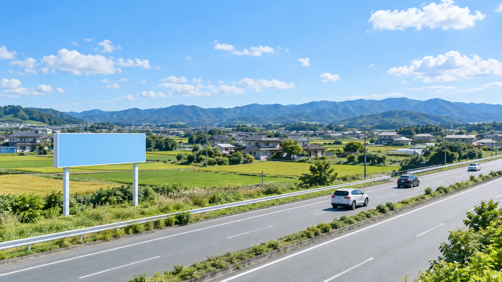 郊外の幹線道路沿いの草地に、2本の柱で支えられた横長の無地の野立て看板が立ち、奥に田畑と低い山並みが広がる様子を、道路の反対側から広く写した写真。看板には文字が無く、車が小さく走っている