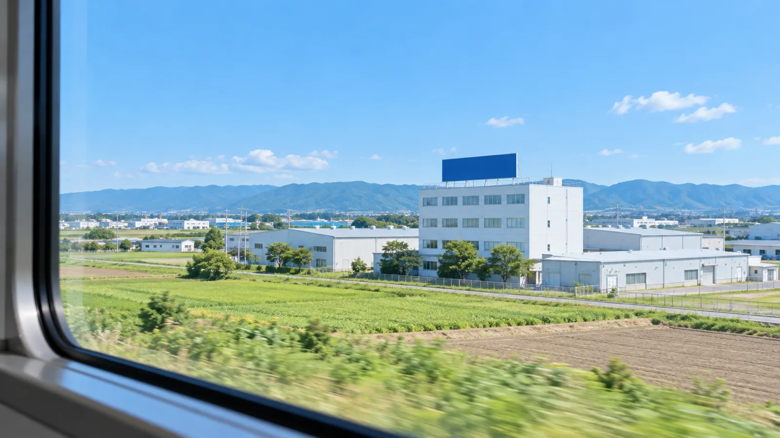 郊外の高架の線路を走る電車の窓から、少し離れた位置に建つ工場の事務所棟を写した写真。窓枠の一部が手前に写り、建物の屋上の端に横長の無地の看板が見える。看板と建物に文字は無く、空は明るい青