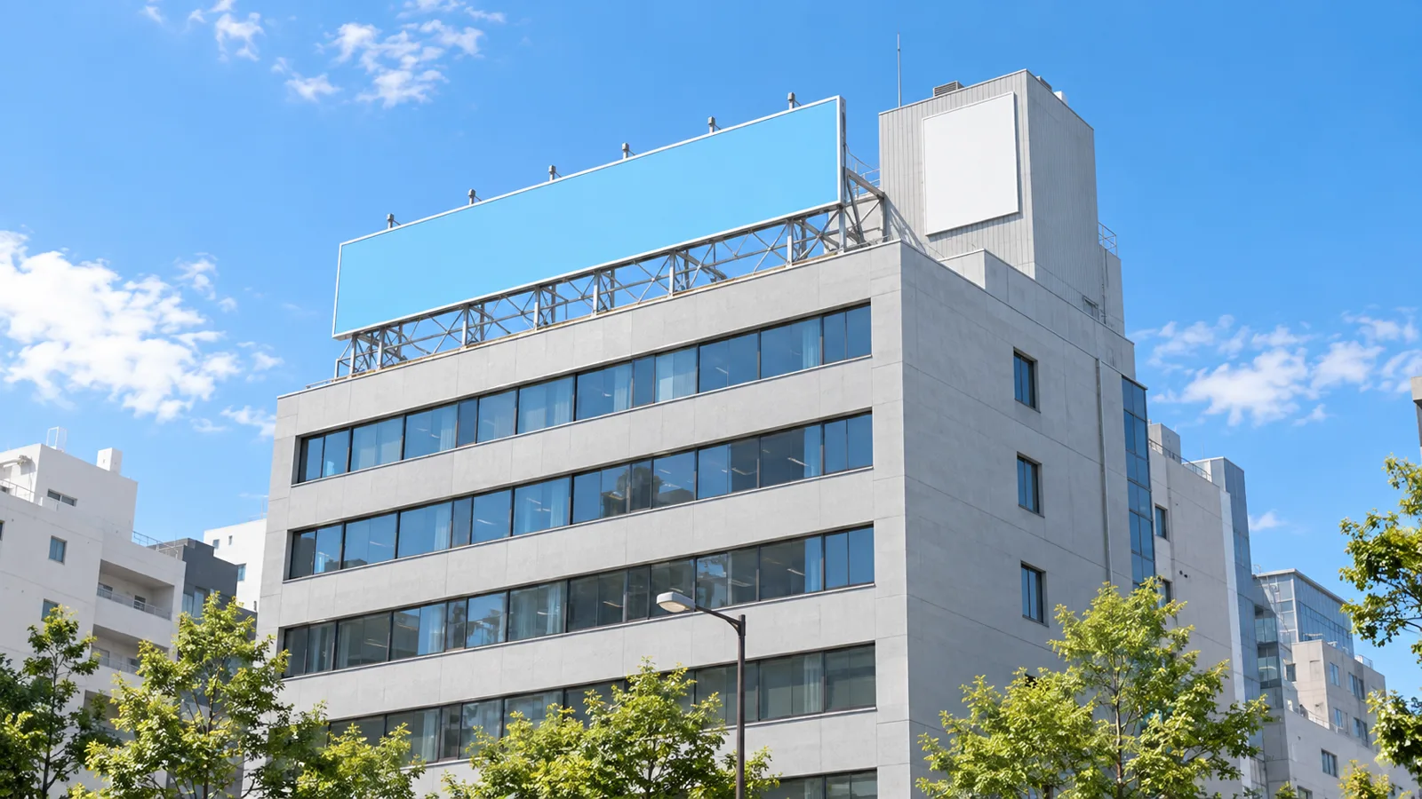 晴れた昼、街の中の7階建てくらいのビルを、少し離れた道路の向こう側から見上げるように写した写真。屋上の端に鉄骨の架台で支えた横長の無地の看板が立ち、その奥の一段高い四角い塔屋の壁にも無地の白い板が付いている。看板と建物にはどこにも文字が無い