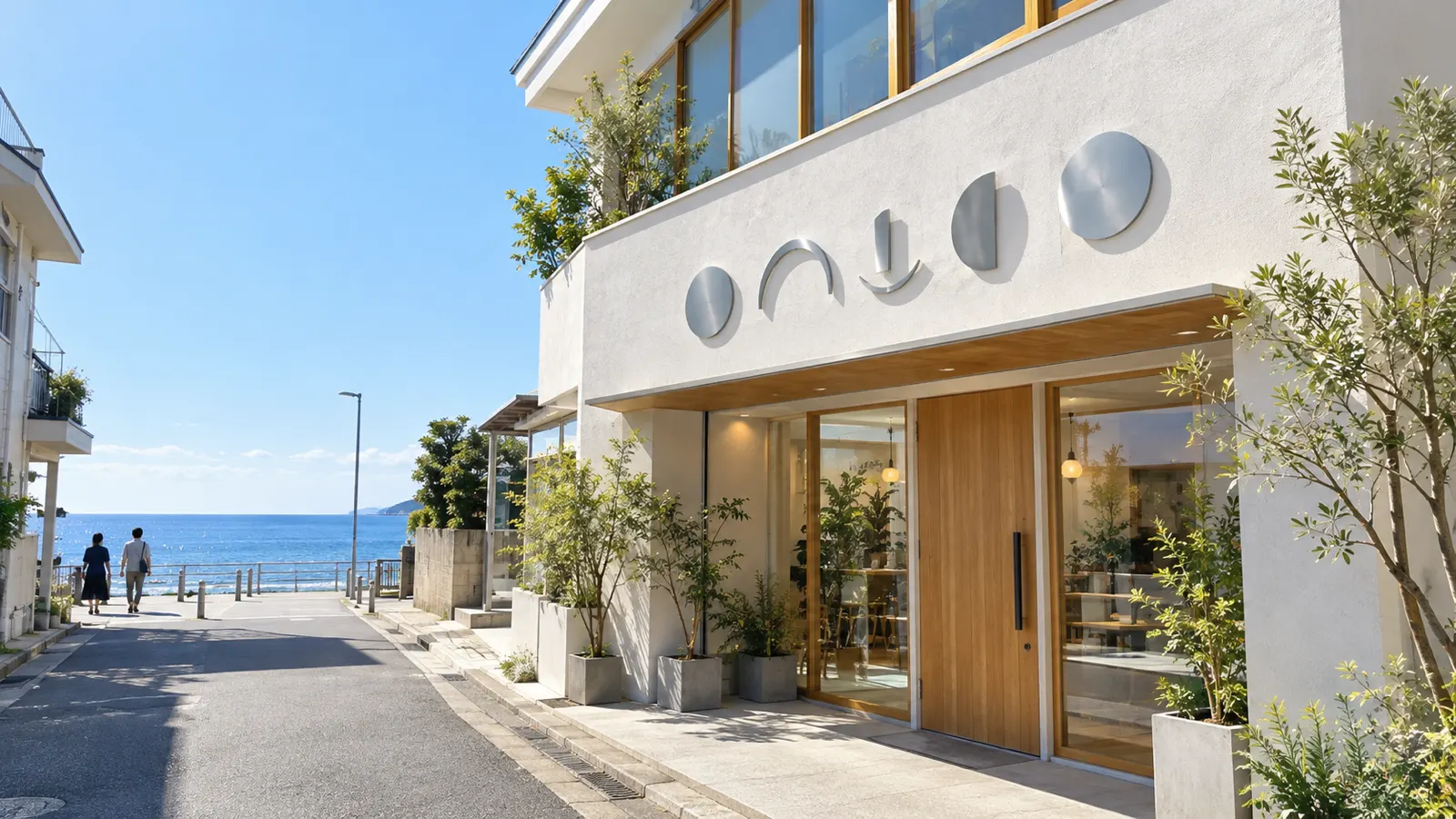 海沿いの通りに面した明るい外壁の店舗を、歩道の少し離れた所から写した広角の写真。外壁にステンレスの丸と弧の形の切り抜きが付き、奥に青い海と晴れた空が見える。人物は小さく通りを歩いている