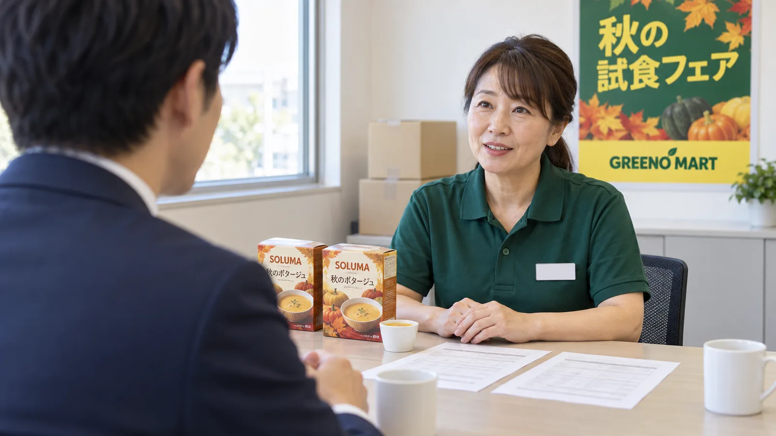 スーパーの事務所のテーブルで、店の販促担当とメーカーの担当者が向かい合って座り、試食に出す商品のサンプルと打ち合わせの紙を前に話している様子を、担当者の肩越しに写した写真