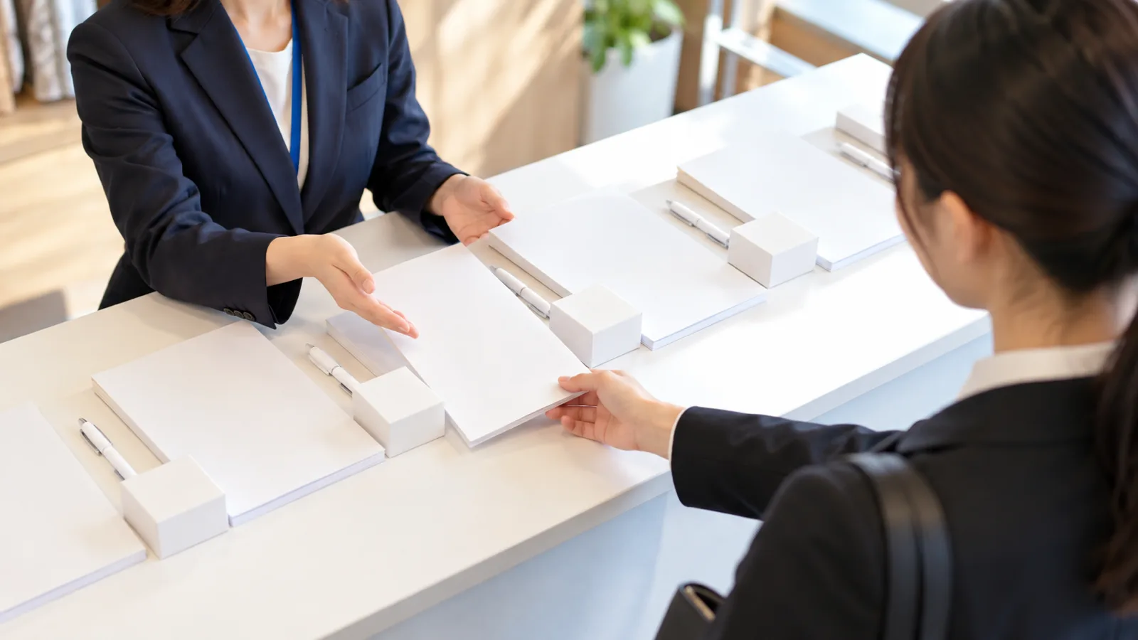 明るい会議室の会社説明会の受付の机に、無地の白い会社案内の冊子と、小さな箱に入った自社の製品のサンプルと、ボールペンを1組ずつ並べ、スーツの学生が1組を受け取ろうとしている様子を、机の斜め上から写した写真。冊子や箱に文字やロゴは無い