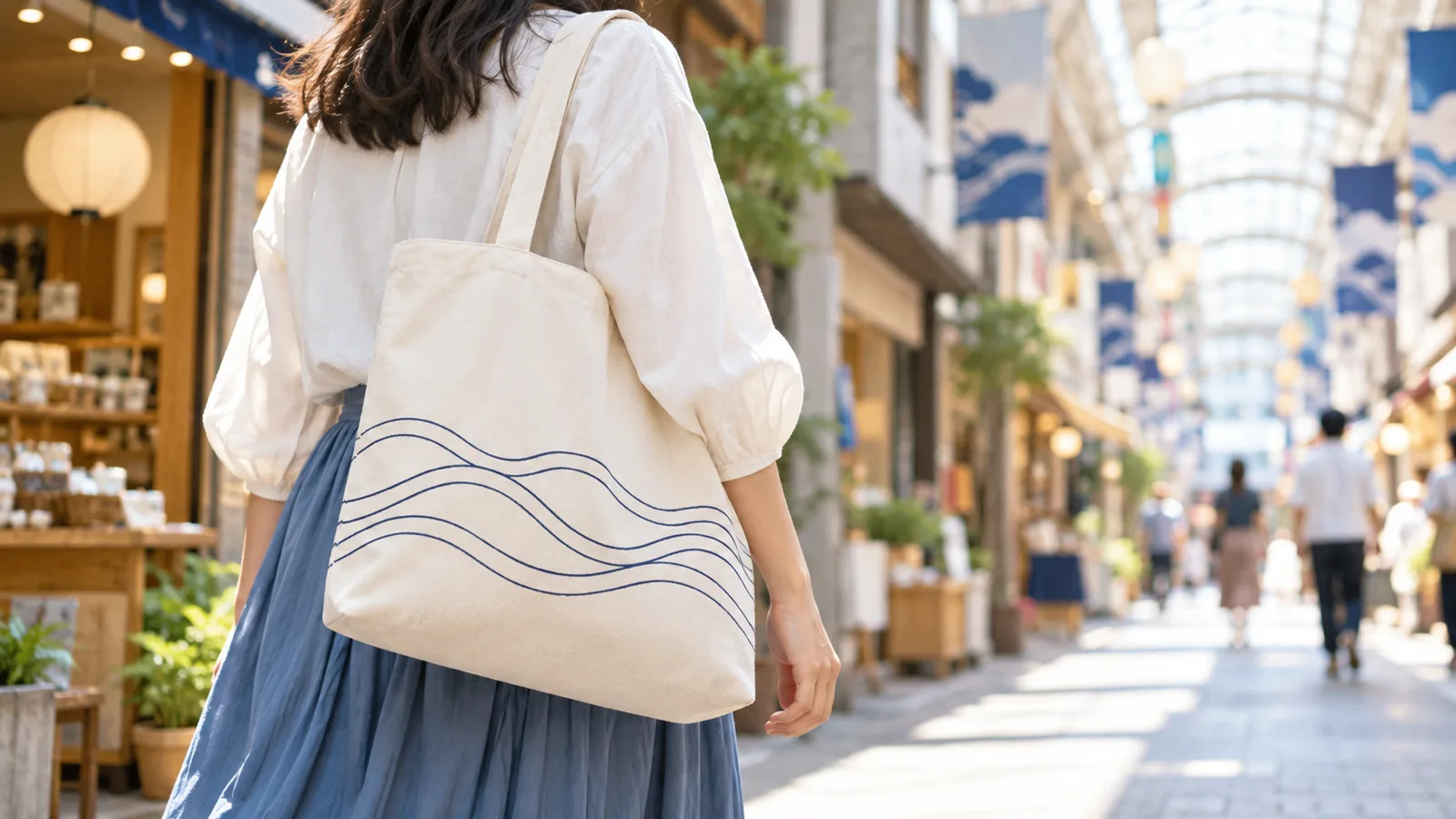 晴れた昼の商店街を歩く女性の肩に掛かった、生成りの布のトートバッグを、斜め後ろの腰の高さから写した写真。バッグには文字の無い控えめな線の模様だけがあり、奥の商店街はやわらかくぼけている